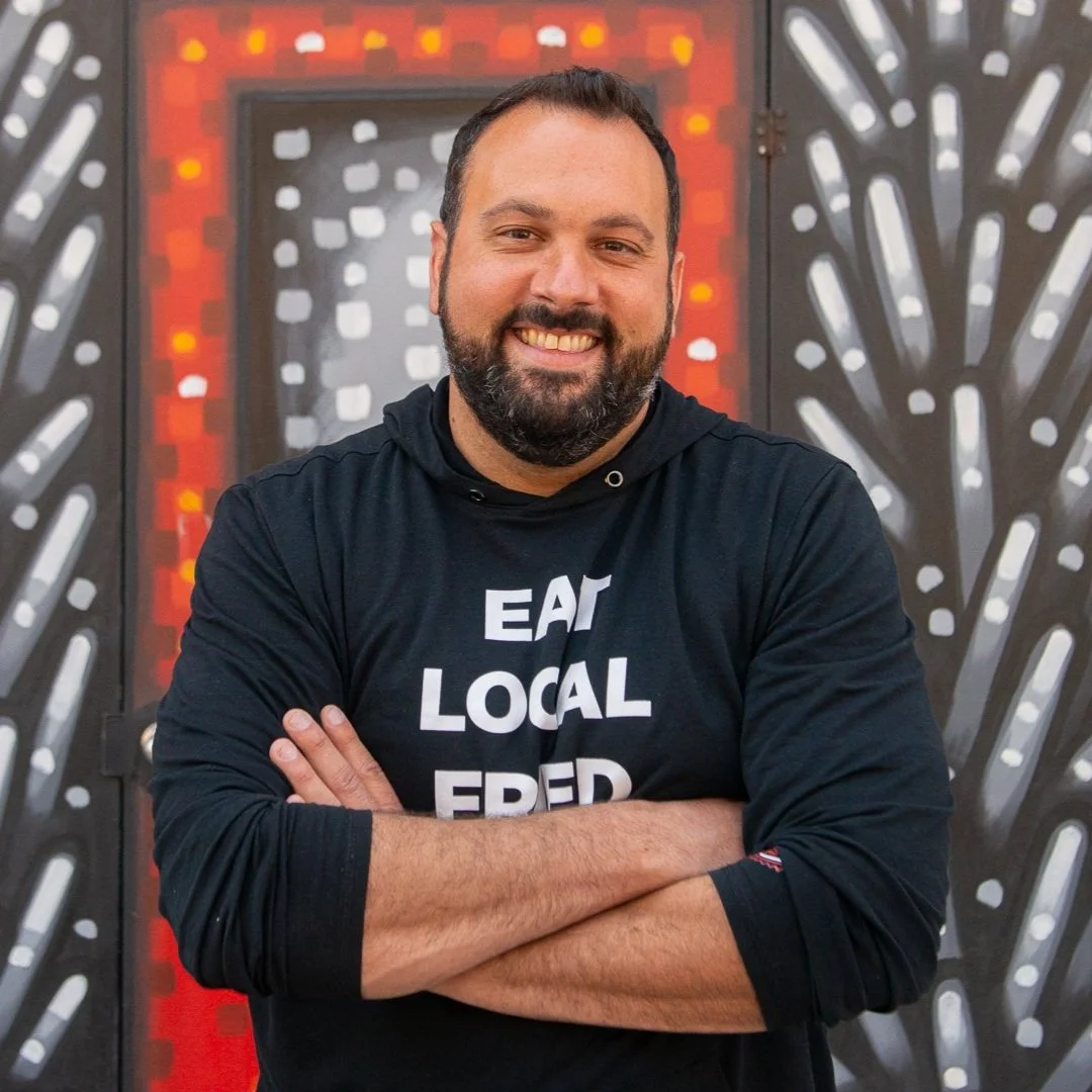 A smiling man with a beard stands in front of a colorful mural, arms crossed. He wears a black hoodie with the words “EAT LOCAL FRED” in bold white letters. The background features geometric black, white, and red patterns.